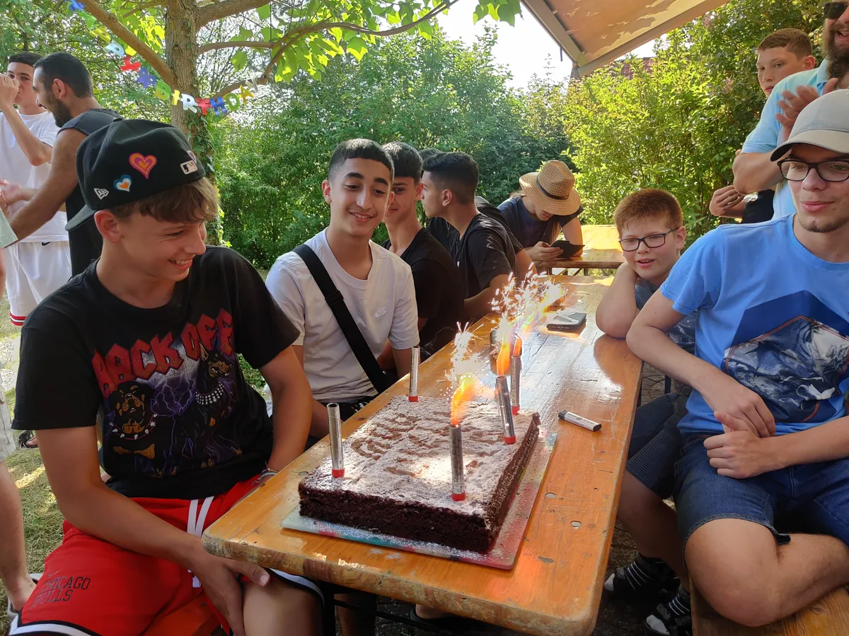Ein Kuchen mit Kerzen als Geburtstagsüberraschung für einen Jugendlichen, der gemeinsam mit anderen auf einer Biertischgarnitur sitzt.