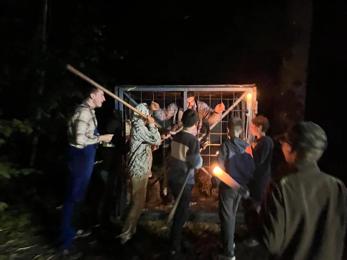 5 Jungs stehen in Dunkelheit mit Fakeln und Stöcken vor einer Gitterbox in der ein Mann (fiktiver Bürgermeister Mai) eingesperrt ist.