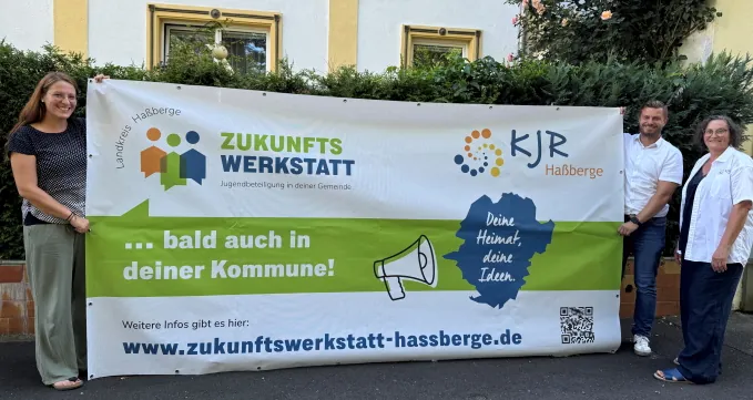 Eva Pfeil (KJR-Geschäftsführerin, Kreisjugendpflegerin), Thomas Wagenhäuser (stellvertretender KJR-Vorsitzender) und Susanne Makowski (KJR-Vorsitzende) halten das Bauzaunbanner für die 2025 erstmals angebotenen Zukunftswerkstätten in die Kamera.