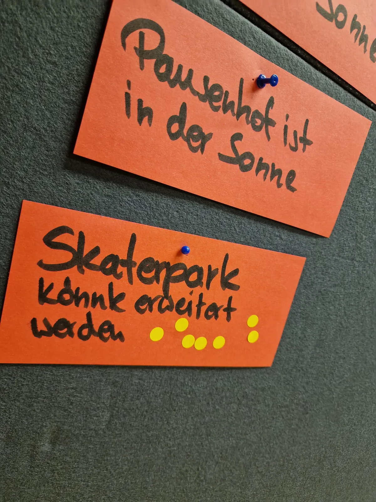 Auf eine grauen Pinnwand sind 2 Moderationskarten gepinnt "Pausenhof ist in der Sonne" und "Skaterpark könnte erweitert werden". Die Skaterpark-Karte hat viele gelbe Votingpunkte.