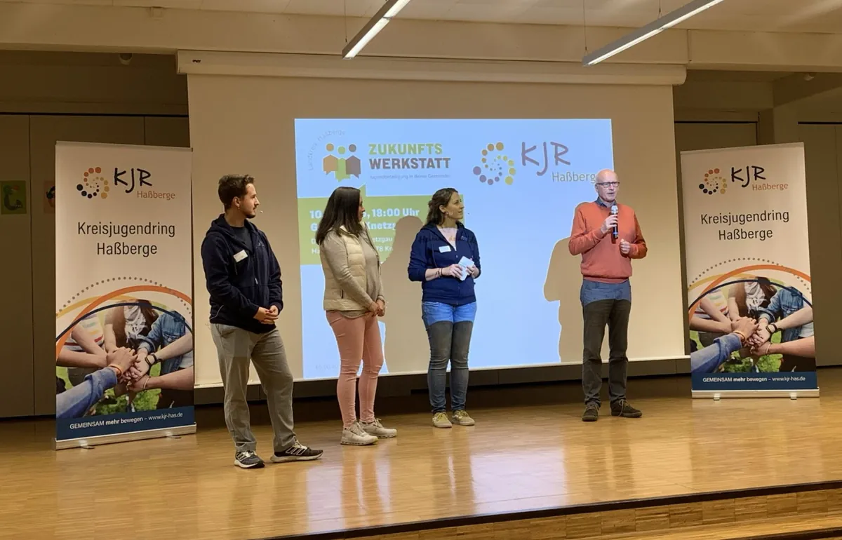 Yannick Reuß, Nina Köberich, Eva Pfeil und Stefan Paulus stehen mit Mikrofonen bei der Zukunftswerkstatt Knetzgau vor einer großen Leinwand und KJR-RollUps auf der Bühne.